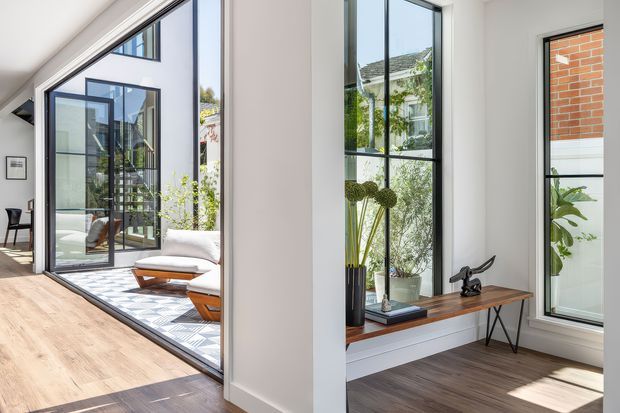 Bright contemporary interior with pale timber flooring, crisp white walls, and extensive black-framed floor-to-ceiling glazing. The view looks through large glazed openings toward a sunlit courtyard or adjoining space, while a built-in timber window seat or console with a small black sculptural object occupies the foreground. A glimpse of a dining or living area appears at left, with indoor plants and restrained furnishings adding a fresh, airy character.