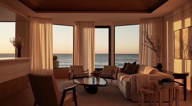 A warm, refined living room overlooking the ocean through expansive floor-to-ceiling glazing. The room features a built-in fireplace or low architectural joinery element, timber ceiling detailing, sheer curtains, a neutral upholstered sofa, sculptural coffee table, timber lounge chairs, and contemporary decorative furnishings. Golden sunset light creates a calm, coastal atmosphere.