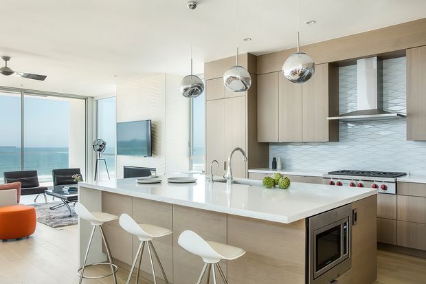 Bright open-plan contemporary kitchen with a large white island, integrated microwave, light timber cabinetry, white subway-tile splashback, stainless-steel rangehood and appliances. Three pendant lights hang above the island, which is paired with white bar stools. A living area and full-height glazing overlooking the sea are visible beyond the kitchen.
