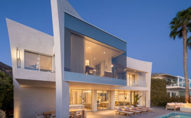 Contemporary multi-level white-rendered residence photographed at dusk, featuring strong geometric volumes, extensive floor-to-ceiling glazing, projecting upper-level glass enclosure, recessed lighting, balconies, outdoor loungers, and a visible swimming pool. Palm trees and neighbouring residences frame the property.