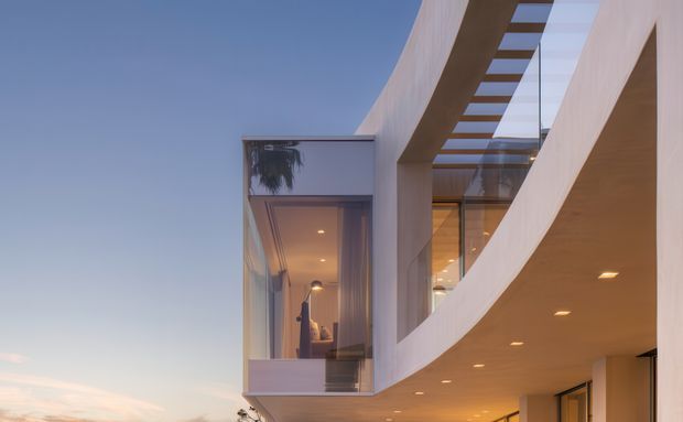 Exterior architectural view at dusk of a contemporary multi-level building with sweeping curved white facade bands, a projecting glazed room, recessed downlighting, and a linear skylight or overhead glazing detail. Warm interior light contrasts with the cool evening sky.
