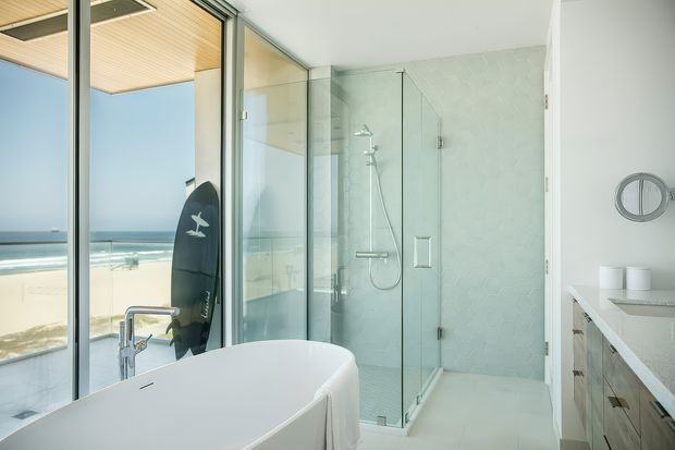Bright contemporary bathroom with a freestanding white bathtub, frameless glass shower enclosure, wall-mounted shower fittings, pale patterned wall tiles, and light timber vanity cabinetry. Full-height glazing opens views toward a sandy beach and ocean, while a surfboard leans beside the window. The room has a clean, airy coastal character.