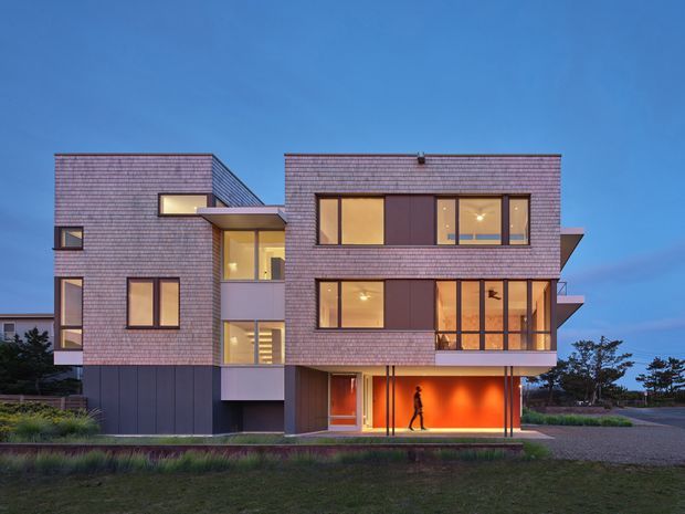 A contemporary multi-storey residential building photographed at dusk, with a rectilinear composition of projecting brick-clad volumes, dark lower-level cladding, extensive warm-lit glazing, balconies, and a recessed illuminated ground-level entry or garage-like opening. The building sits in a landscaped suburban or coastal-looking setting, viewed from the front exterior.