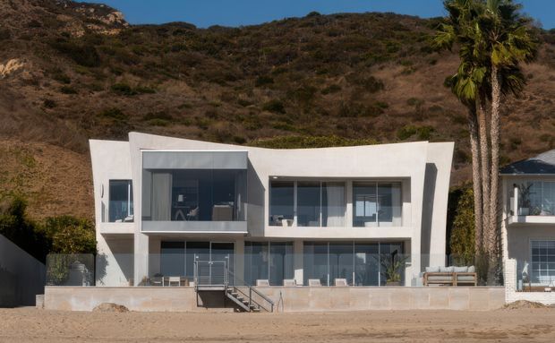 A contemporary two-storey white coastal house set directly against a dry, shrub-covered hillside. The facade features broad glazed openings, projecting upper-level volumes, flat rooflines, balconies, and a central exterior stair leading to the beach-level frontage. A tall palm and a neighbouring residence are visible at the right edge.