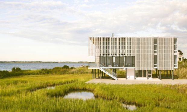 Exterior architectural photograph of a contemporary elevated coastal house set beside a marshy waterfront. The rectangular multi-level building has extensive glazing, a light vertical screen or louvred facade, slender supports, and an external stair leading to the raised living level. Reeds, tidal pools, and calm water surround the structure under a bright partly cloudy sky.