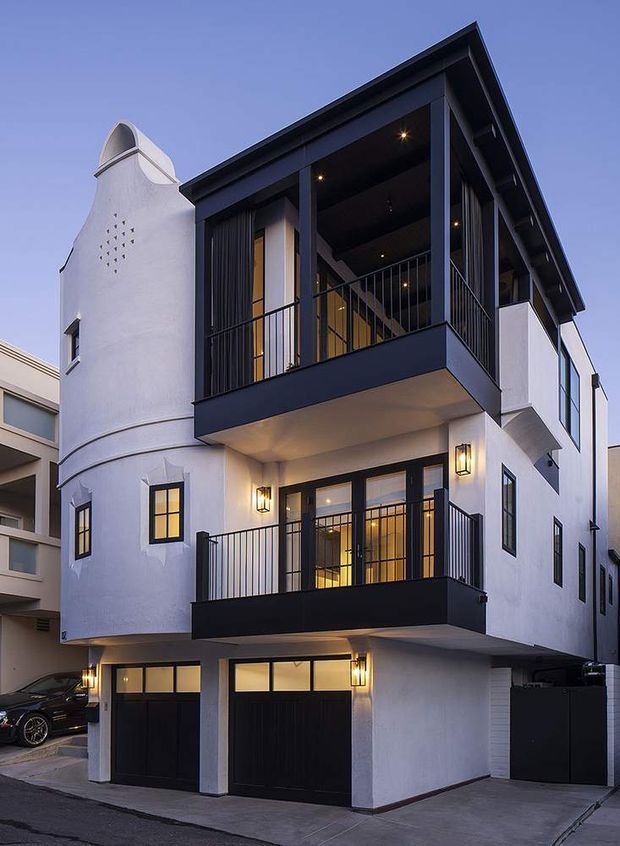 Exterior architectural photograph of a narrow multi-storey urban residence at dusk. The building has a white stucco facade, strong black-framed glazing and railings, two projecting balconies, ground-level garage doors, and a distinctive curved tower-like side volume with small perforated openings and a sculptural arched roof detail. Warm wall sconces and interior lighting contrast with the dark blue-black exterior trim.