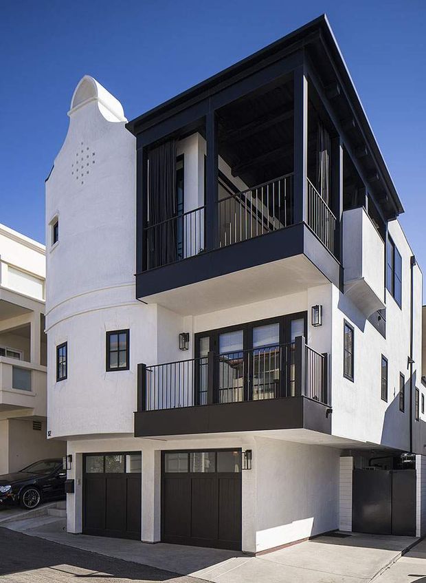 Exterior street view of a multi-storey contemporary residential building with white rendered walls, strong black trim, stacked balconies, open upper-level terrace, black metal railings, and dark garage doors. A distinctive curved parapet detail appears on the adjoining facade, while the main building has a flat, box-like roof form.