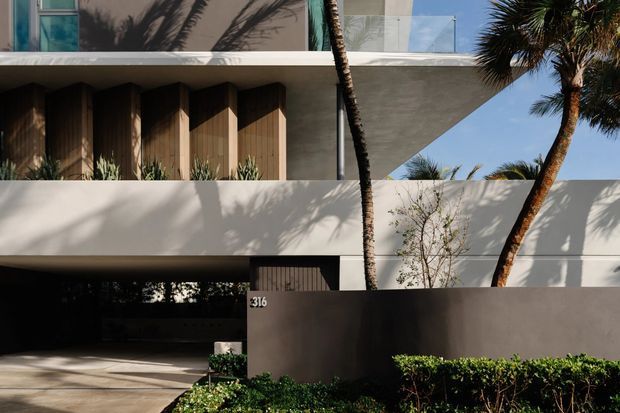Contemporary multi-storey residential facade with crisp white horizontal volumes, a projecting upper balcony with glass balustrade, timber vertical screening at the entry, and a dark rendered boundary wall. Tropical palms and landscaped hedging frame the street-facing composition, with a partially concealed carport or garage opening at ground level.