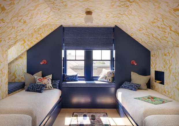 A compact attic-style twin bedroom with two built-in single beds facing each other. The room features deep navy-blue built-in bed bases and wall panels, patterned bedding and cushions, a central window with a navy Roman blind, and sloped walls and ceiling covered in a bold cream-and-gold botanical pattern. Small red wall lights and a colourful central rug add playful accents.
