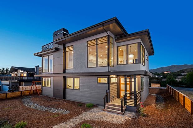 Exterior view of a contemporary multi-level house at dusk, featuring a dark grey and light-rendered facade, extensive floor-to-ceiling glazing, projecting roof forms, a rooftop chimney or vent, balcony railings, and a timber-and-metal front entry stair. The home sits on a landscaped gravel lot in a suburban or semi-rural setting with hills visible in the background.