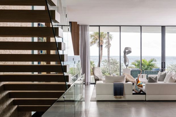 Contemporary open-plan living room viewed from a dramatic floating staircase. The room has a large white sectional sofa, sculptural decor, floor-to-ceiling glazing, and an expansive ocean-facing outlook with palms beyond. A glass balustrade and dark timber stair treads create a strong architectural contrast with the light, airy interior.