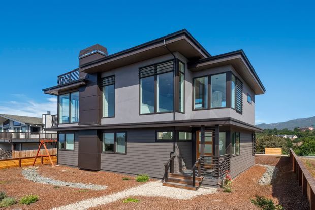 Exterior view of a contemporary two-storey house with a low-pitched roof, dark horizontal cladding, smooth rendered upper walls, extensive floor-to-ceiling glazing, projecting upper-level volumes, metal balcony railings, and a small entry stair. The home sits on a landscaped gravel and planting site with neighbouring houses and hills visible in the background.