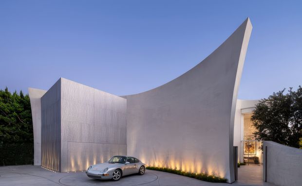 Exterior view of a sculptural contemporary residence or entry pavilion, dominated by a tall curved pale concrete wall that rises into a sharp pointed fin. Ribbed vertical cladding forms an adjoining facade, with a partially visible illuminated glazed entry behind a side wall. A paved forecourt and parked sports car sit in front, with landscaping at the edges.
