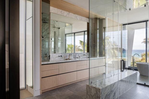 A luxurious contemporary bathroom with a double vanity, pale timber-look cabinetry, expansive mirrors, marble-effect benchtops and feature wall surfaces, and a frameless glass shower enclosure. A freestanding bathtub is visible at the far right, with large windows overlooking palm trees and the ocean. The room has a refined, bright, resort-like atmosphere.
