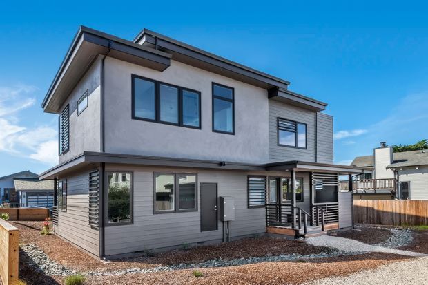 Exterior view of a contemporary two-storey residential house with a mixed light rendered and horizontal grey cladding facade. The home has dark-framed rectangular windows, projecting roof planes, a covered entry porch, and landscaped gravel surrounds. Neighboring homes are visible in the background under a clear blue sky.