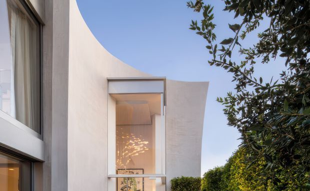 Exterior architectural view of a sculptural contemporary building with smooth pale rendered walls, a dramatically curved roof or parapet edge, and tall glazed openings. Warm interior lighting and a pendant or feature light are visible through the central glazing, while mature landscaping frames the right side of the composition.