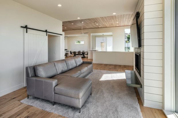 Bright open-plan living room with a large grey sectional sofa and ottoman on a textured rug, facing a built-in fireplace set within white horizontal wall panelling. The room includes warm timber flooring, a timber-lined ceiling over the rear area, a white kitchen island, dining area, sliding barn door, recessed lighting, and large glazed openings.