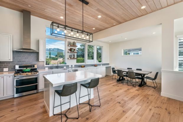 Bright open-plan kitchen and dining area with white shaker-style cabinetry, a large white island, stainless steel range and hood, grey tiled splashback, and timber flooring. A timber-lined ceiling and exposed black-framed pendant light add warmth and industrial character. Large windows overlook a green, hilly setting.