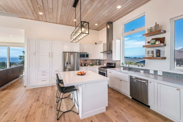Bright coastal-style kitchen with a large white island, integrated seating, extensive white cabinetry, stainless-steel appliances, and a tiled backsplash. A warm timber-plank ceiling and timber flooring contrast with the white fit-out. Large windows and an adjoining opening provide views toward the ocean and coastline. A black linear pendant light and dark rangehood add contrast.