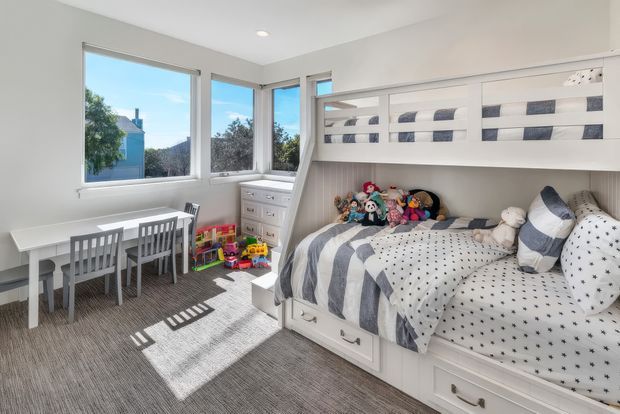 Bright, compact children's bedroom with a built-in bunk bed and lower trundle-style storage drawers, coordinated white and navy striped and patterned bedding, and a collection of soft toys. A small white table with grey chairs sits beside large corner windows, with a dresser and children's toys near the bed. The room has white walls, grey carpet, abundant natural light, and a clean, playful fitted design.