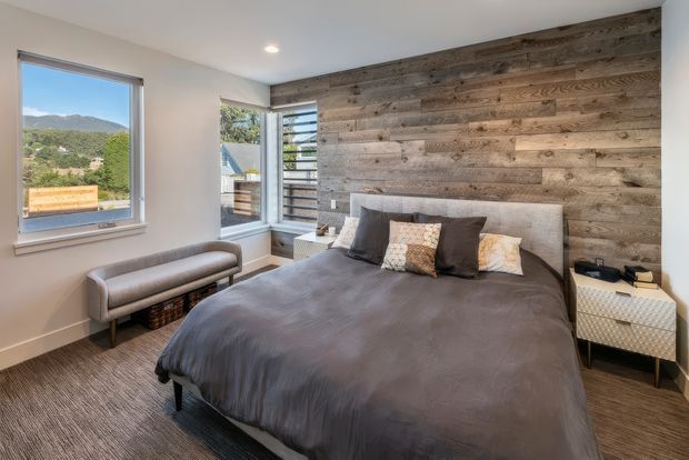 A contemporary bedroom with a large bed dressed in dark grey bedding, a light upholstered headboard, and layered neutral cushions. The room features a full-height reclaimed timber feature wall behind the bed, light painted walls, grey carpet, a compact upholstered bench, and a patterned bedside table. Windows and a glazed corner opening provide views toward a green, mountainous landscape.