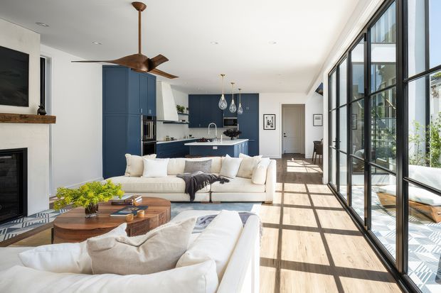Bright open-plan living room and kitchen with expansive black-framed glazing along one side, a white sectional sofa arrangement, a rounded timber coffee table, and a contemporary navy-blue kitchen with island. A linear fireplace and wall-mounted television sit on the left, while sculptural pendant lights and a timber ceiling fan add focal details. Strong daylight creates a warm, airy atmosphere and highlights the timber-toned floor.