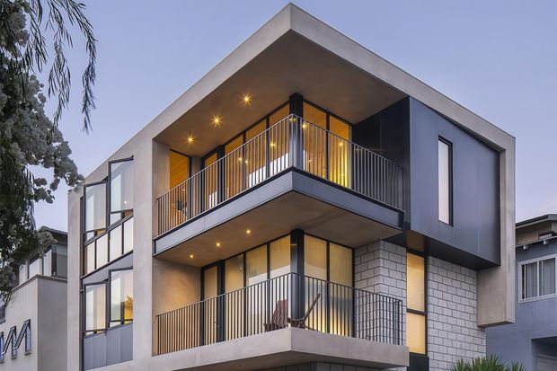 A contemporary multi-storey residential building photographed from the street at dusk. The facade combines smooth light rendered surfaces, dark charcoal cladding, extensive glazing, and a pale masonry feature wall. Two projecting balconies have slim black metal balustrades and recessed warm lighting beneath the roof overhangs. The design has strong geometric massing, deep square openings, and floor-to-ceiling windows.
