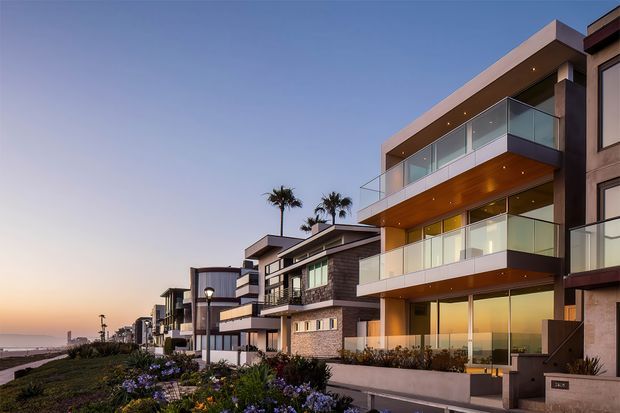 Twilight exterior view of a row of contemporary multi-storey coastal residences lining a landscaped promenade. The foreground home features broad projecting balconies with clear glass balustrades, warm timber soffits, large glazed openings, and a mixed dark and light facade. Adjacent homes continue into the distance, with palm trees, planting, and a visible waterfront or beach edge.