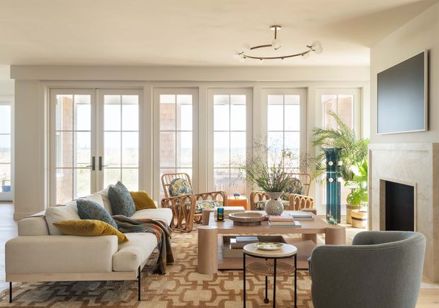 Bright, refined living room with a large cream sectional sofa, patterned area rug, woven rattan accent chairs, a wood-toned rectangular coffee table, and a grey upholstered lounge chair. Tall French doors with divided glazing line the rear wall, bringing in abundant natural light. A pale stone or marble fireplace with a wall-mounted television forms the right-side feature, while indoor palms and decorative furnishings add a relaxed organic touch. The room has a warm neutral palette with mustard and blue-green accents.