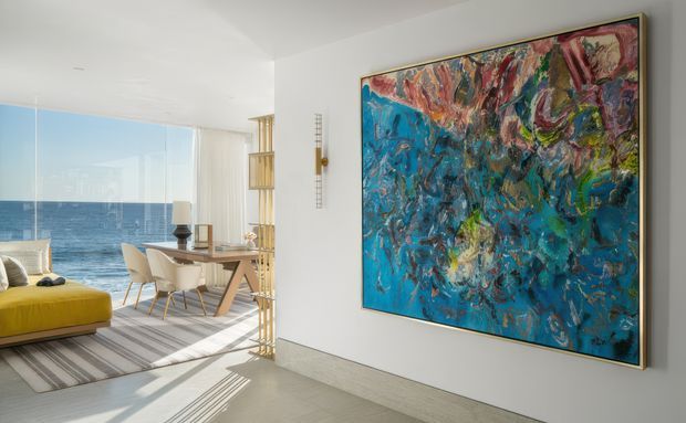 Bright contemporary apartment interior with a large abstract artwork dominating a white wall in the foreground. Beyond the open passage is an open-plan living and dining area with floor-to-ceiling glazing, an ocean view, a yellow sofa, a pale dining table and chairs, and strong daylight casting linear shadows across the pale floor and striped rug. Vertical timber or brass-toned screening separates the dining area from the circulation space.