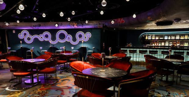 A dark, theatrical cocktail bar or lounge with curved banquette seating, small round tables, red and orange upholstered chairs, a backlit bottle display, and a long illuminated bar counter. A prominent purple neon artwork spans the rear wall, while spherical pendant lights and patterned flooring add a retro-futuristic atmosphere.
