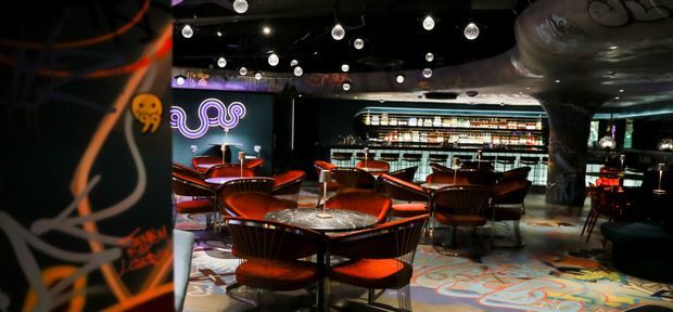 A dimly lit, theatrically designed cocktail bar or lounge with circular tables, curved red-orange upholstered chairs, a long illuminated bottle display and bar counter at the rear, and a highly patterned carpet. Black ceilings feature numerous hanging globe lights, while sculptural ceiling and column elements, neon artwork, and bold graphic surfaces create an immersive nightlife atmosphere.