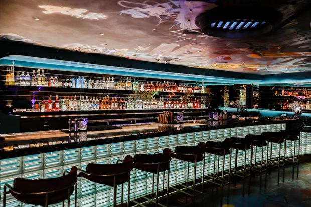 A dramatic, low-lit cocktail bar interior with a long illuminated bar counter, rows of liquor bottles displayed on backlit shelving, dark bar stools, reflective black surfaces, and a highly reflective sculptural ceiling with metallic and graphic details. The atmosphere is luxurious, moody, and nightlife-oriented.