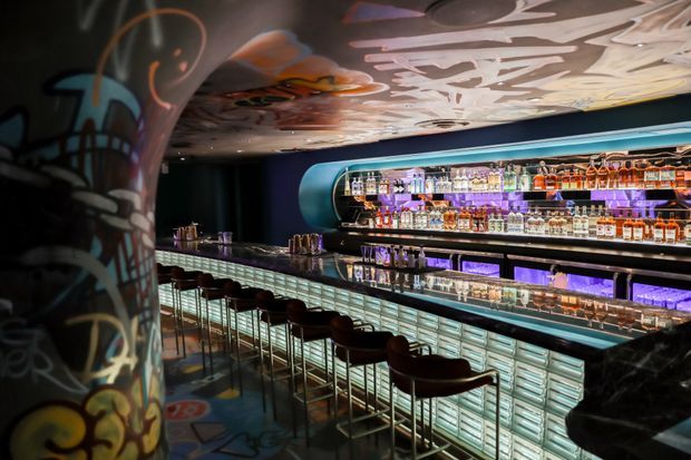 A dark, highly stylised bar interior with a long illuminated drinks counter, extensive backlit bottle shelving, reflective surfaces, and a row of dark bar stools. The space features colourful graffiti-style artwork on a large column and across the ceiling, with purple and turquoise accent lighting creating a dramatic nightlife atmosphere.