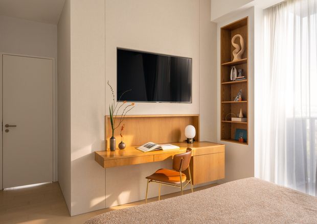 A warm, softly lit bedroom featuring a built-in timber entertainment and study wall. The joinery includes a wall-mounted television, floating desk, drawers, open display shelving, decorative objects, and a compact mid-century-style chair. A bed occupies the foreground, while sheer curtains provide filtered daylight beside the built-in cabinetry. The room has a restrained, contemporary look with warm timber and beige finishes.