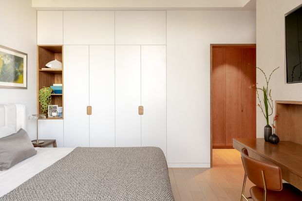 Bright, compact bedroom with a neatly made bed, warm timber flooring, full-height white built-in wardrobes, and an integrated timber desk and shelving unit. A timber door, bedside table, framed artwork, soft neutral bedding, and simple decorative items create a calm, contemporary interior.