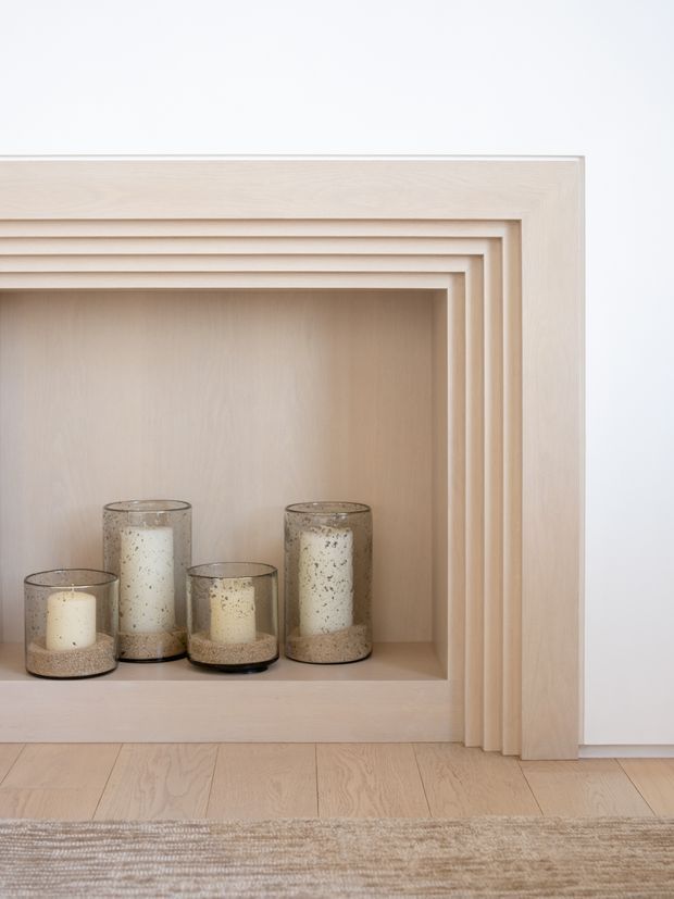 Close-up of a minimalist built-in timber feature recess with multiple stepped rectangular surrounds, displaying several glass candle holders. The pale wood joinery continues onto a light timber floor, with a neutral textured rug in the foreground and a plain white wall.