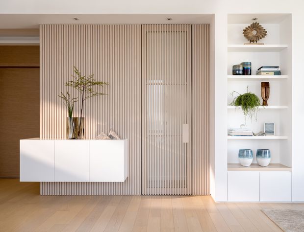 Bright contemporary built-in living area featuring a full-height fluted timber feature wall and concealed slatted door, a floating white storage cabinet, and integrated white display shelving with curated decor. Pale timber flooring and warm neutral finishes create a refined, airy interior.