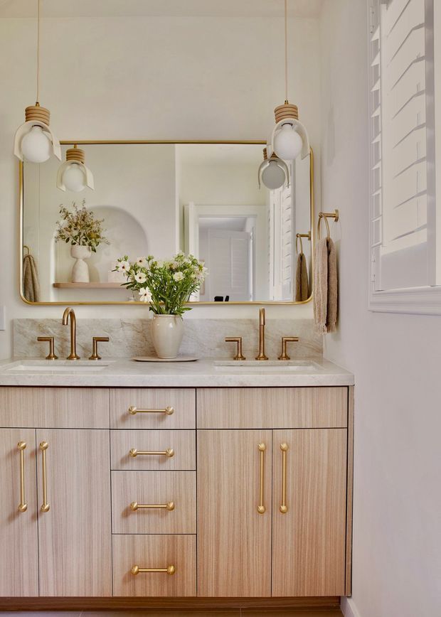 A refined bathroom vanity with a double-basin marble countertop, pale vertical-grain timber cabinetry, polished brass tapware and handles, a large rounded-corner brass-framed mirror, globe pendant lights, and white plantation shutters. Soft floral styling adds warmth to the otherwise light, elegant interior.