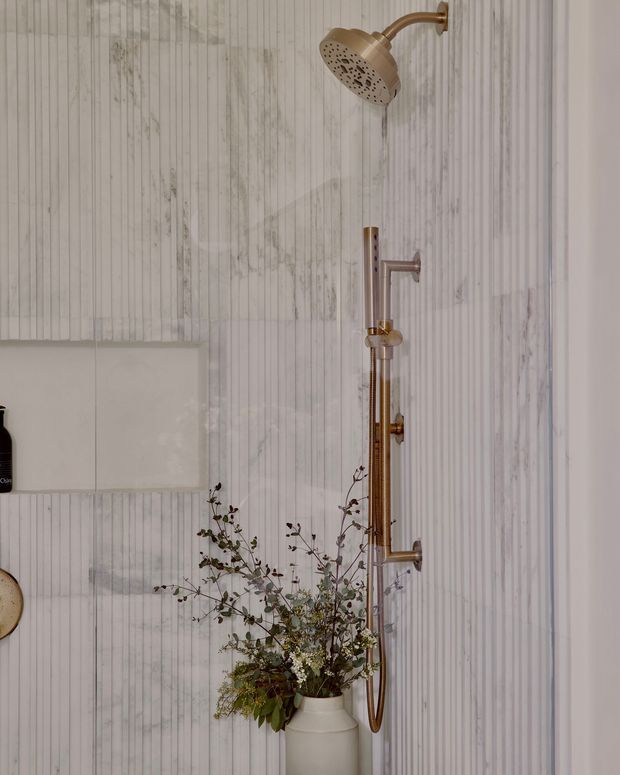 Close-up view of a bathroom shower enclosure featuring a warm brass exposed shower system with overhead showerhead and handheld rail, set against vertically fluted pale stone or tile wall panels. A white vessel containing leafy branches sits below, with minimal wall-mounted accessories at the side.