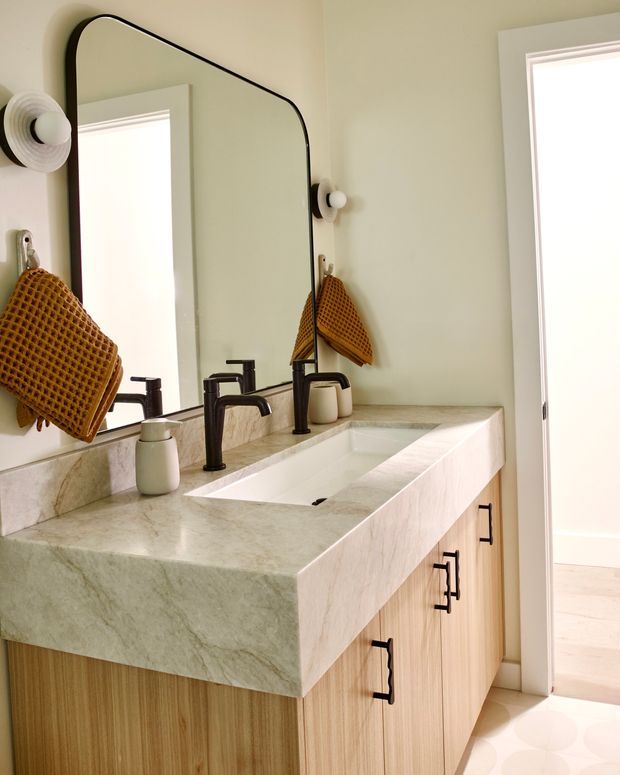 Bright bathroom vanity with a substantial pale veined stone countertop, integrated rectangular sink, twin dark metal faucets, and light timber cabinetry with black pulls. A large rounded-corner black-framed mirror spans the vanity, with sculptural wall sconces and a warm ochre waffle-textured hand towel. The room opens toward a bright adjacent area through a doorway.