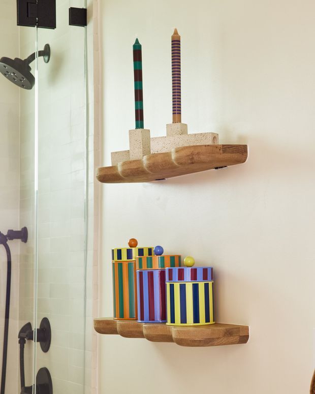 A colourful, playful bathroom vignette showing a glass-enclosed shower on the left and two floating wooden shelves mounted on a light wall. The upper shelf holds a sculptural wooden rack with striped candle-like objects, while the lower shelf displays several brightly striped cylindrical containers. The room has a warm, eclectic character with simple contemporary bathroom fixtures.