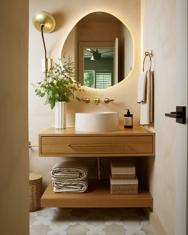 Warm, compact bathroom vanity with a floating natural wood drawer unit and open lower shelf, a white vessel basin, brushed brass wall-mounted tapware, and a large rounded organic-shaped mirror with integrated backlighting. Neutral plaster walls, patterned light stone or ceramic floor tiles, brass lighting and hardware, folded towels, and botanical styling create a refined contemporary interior.