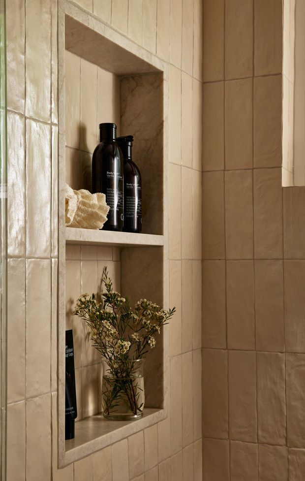Close-up of a warm, earthy bathroom wall clad in handmade-look beige ceramic tiles, featuring recessed tiled niches with stone-like shelves holding dark toiletry bottles, a folded towel, and a small vase of dried greenery. The space has a refined, softly lit, natural spa-like atmosphere.