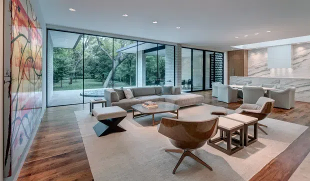 A spacious, open-plan contemporary living room with floor-to-ceiling glazing overlooking greenery. The room features warm timber flooring, a large pale area rug, a sectional sofa, sculptural lounge chairs, nesting tables, recessed ceiling lights, and a sleek kitchen or bar area with marble-look wall surfaces and timber cabinetry in the background. A large colourful abstract artwork forms a bold feature wall.
