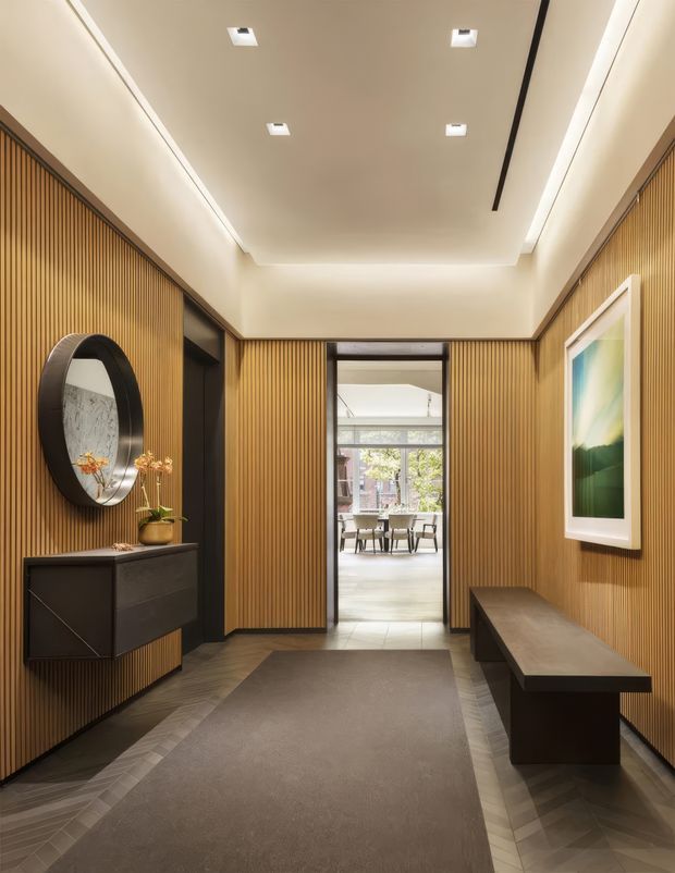 A refined residential entry hall with full-height warm timber slat wall panelling, a neutral recessed ceiling with integrated linear and downlight illumination, dark built-in cabinetry, a large circular mirror, framed artwork, and a substantial dark bench. A herringbone timber floor border surrounds a large taupe rug, while a glazed opening leads to a bright dining area beyond.