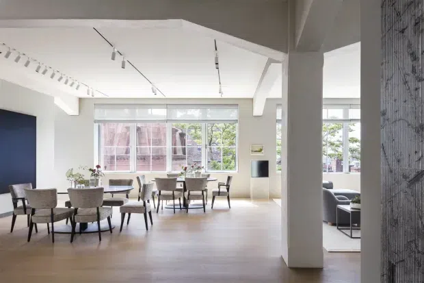 Bright open-plan interior with a dining area in the foreground and a partially visible living area beyond a substantial structural column. The space features pale timber flooring, white walls and exposed white-painted beams, large industrial-style windows, and linear track lighting. Several dining tables and upholstered chairs create a refined, contemporary hospitality or residential entertaining setting.
