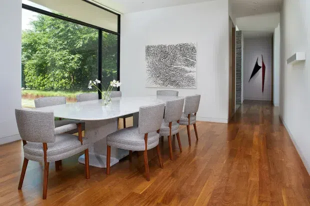 Bright contemporary dining area with a long oval light-toned dining table and upholstered timber-leg chairs, set against white walls and warm timber flooring. A large black-framed glazed opening provides views to greenery, while a minimalist artwork and hallway extend into the background.