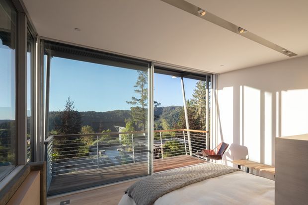 Bright contemporary bedroom with expansive floor-to-ceiling sliding glass doors opening to a timber balcony. The room has pale walls, a light upholstered bed, a textured grey throw, a modern lounge chair, recessed track lighting, and broad views toward a wooded hillside. Slim dark window frames and a built-in light-toned bedside or storage element contribute to the minimalist design.