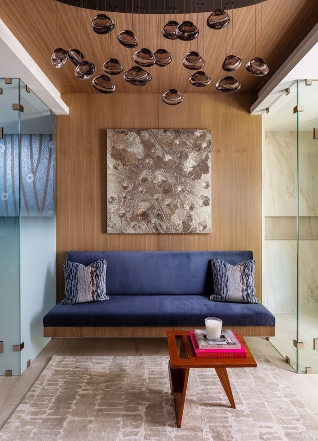 A refined interior seating area with a deep navy upholstered built-in sofa set against warm vertical timber wall panelling. A large abstract artwork hangs above the sofa, while a sculptural cluster of dark glass pendant lights hangs from the timber-lined ceiling. Transparent glass partitions flank the space, and a small mid-century-style timber coffee table sits on a pale patterned rug with books and a cup.